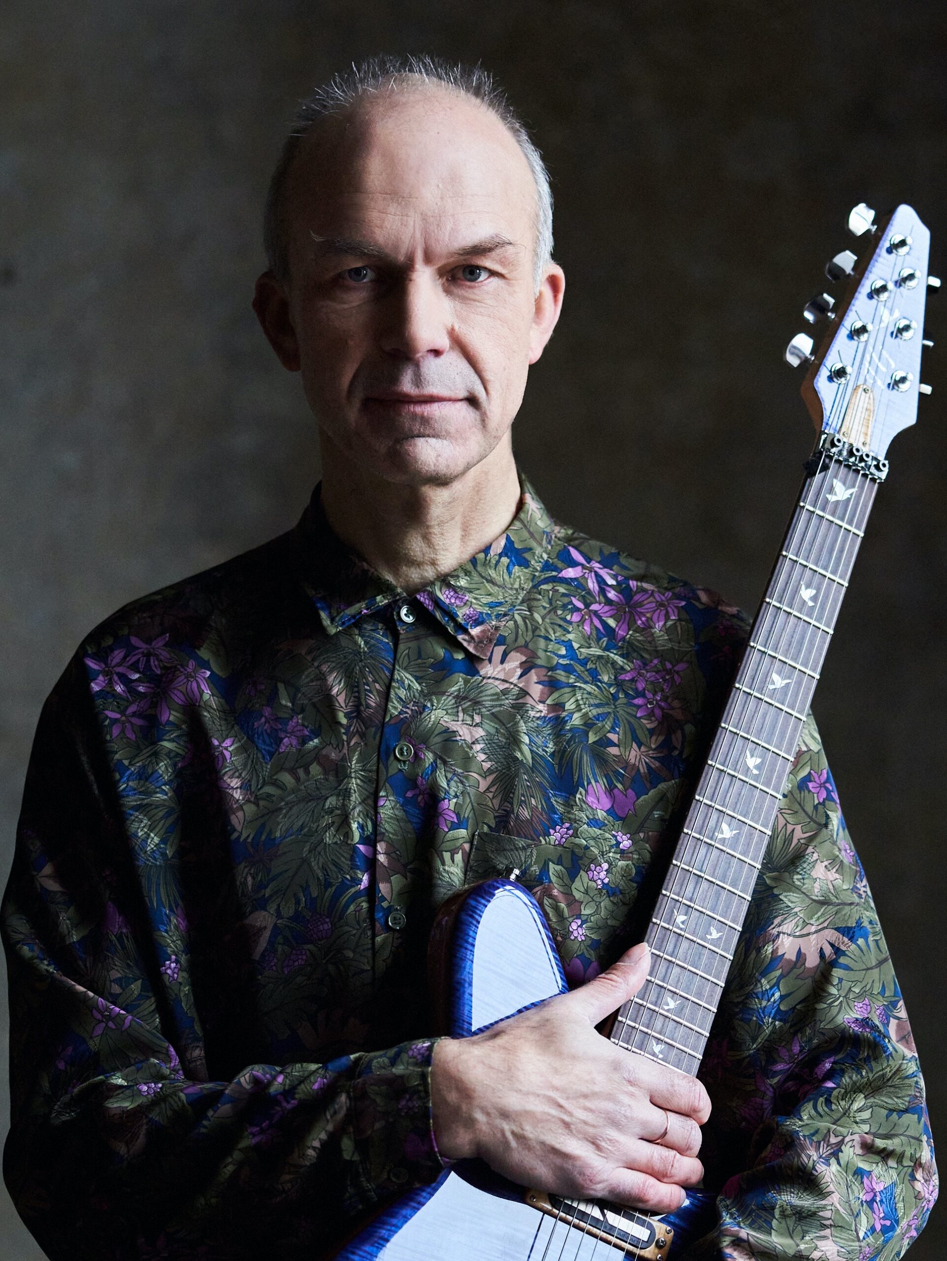 Man in a floral shirt holds a blue guitar in a dimly lit room, staring seriously at the camera. Neutral expression, plain background.